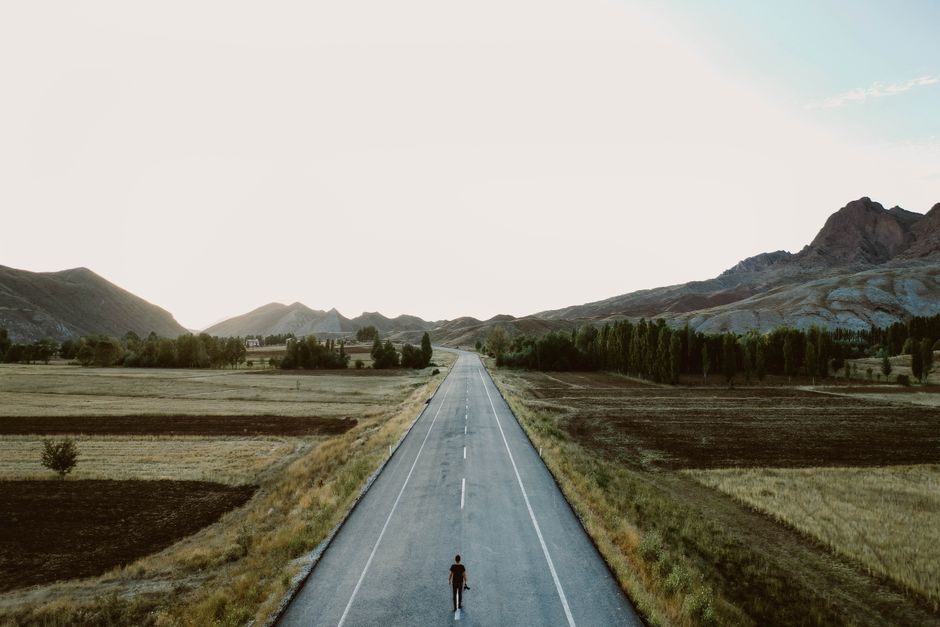 A lone person walks on a rural highway surrounded by fields and mountains under a vast sky.